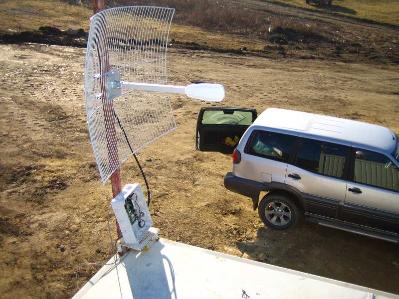 A parabolic grid antenna on a pole beside a silver 4x4 with its boot open, on bare winter ground