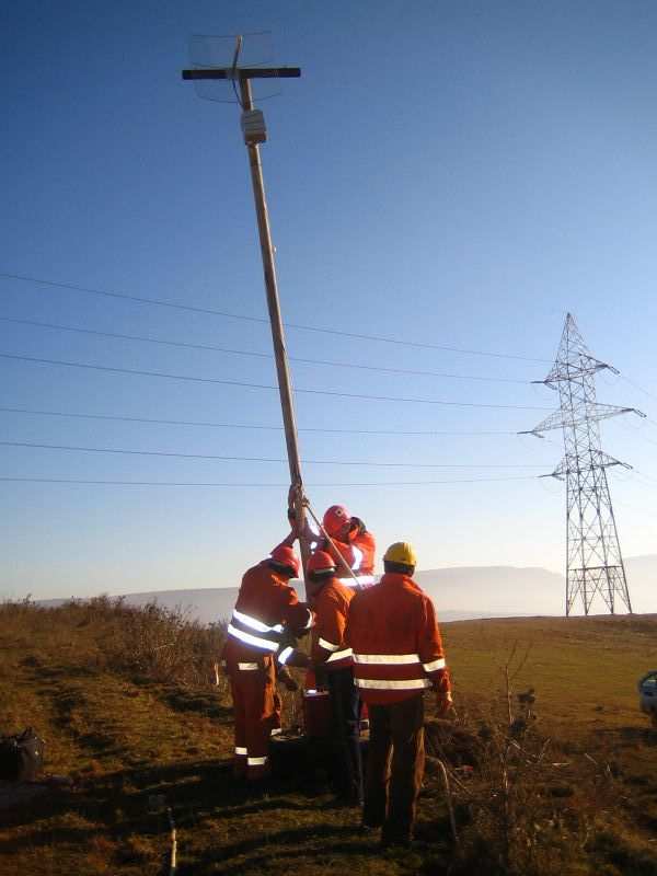 Four workers in orange overalls and hard hats raising a wooden mast with an antenna, a high-voltage pylon behind them under a clear blue sky