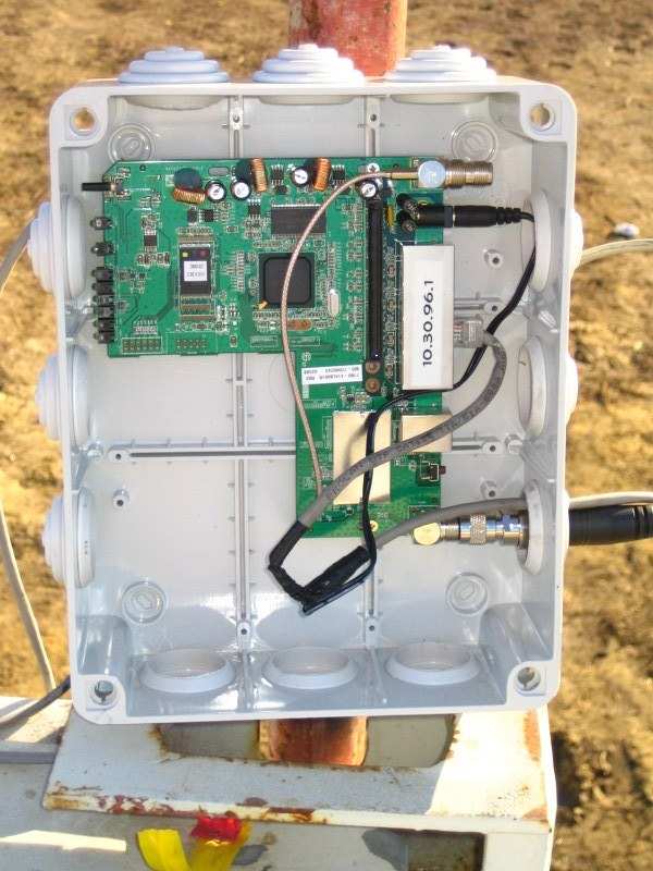 A WRT54GL router board mounted inside a white polycarbonate weatherproof box on a pole in a muddy field, labelled with the IP 10.30.96.1